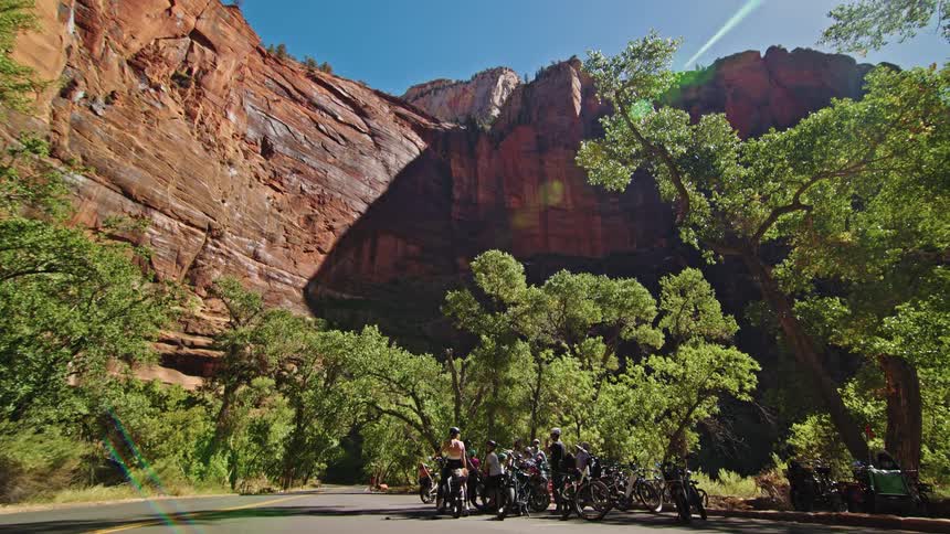 A low angle wide view shows a group of cyclists taking a break near their bikes on the side of a road with towering red sandstone cliffs and vibrant green trees surrounding them on a sunny day