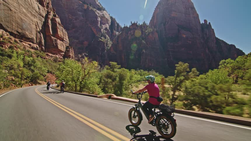 A woman wearing a pink shirt and a helmet rides a thick tire electric bike down a sunny road following other cyclists through a valley surrounded by immense red rock formations and green trees