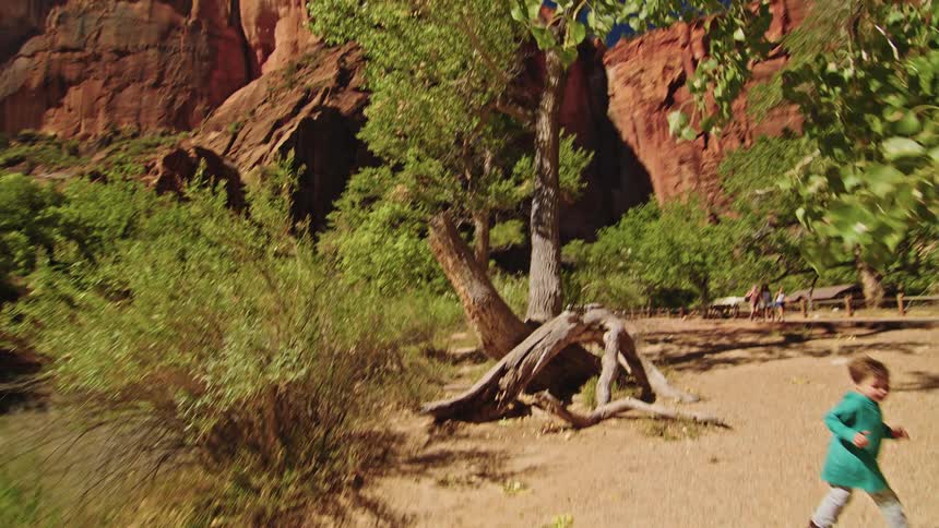 A small child wearing a blue shirt walks along a sandy riverbank past green trees with towering red sandstone cliffs in the background under a bright blue sky