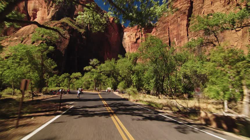 Group of cyclists rides along a road surrounded by trees and reddish cliffs