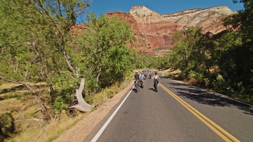 A large group of cyclists wearing helmets are riding bicycles down a paved road surrounded by green trees and majestic red sandstone cliffs on a bright sunny day