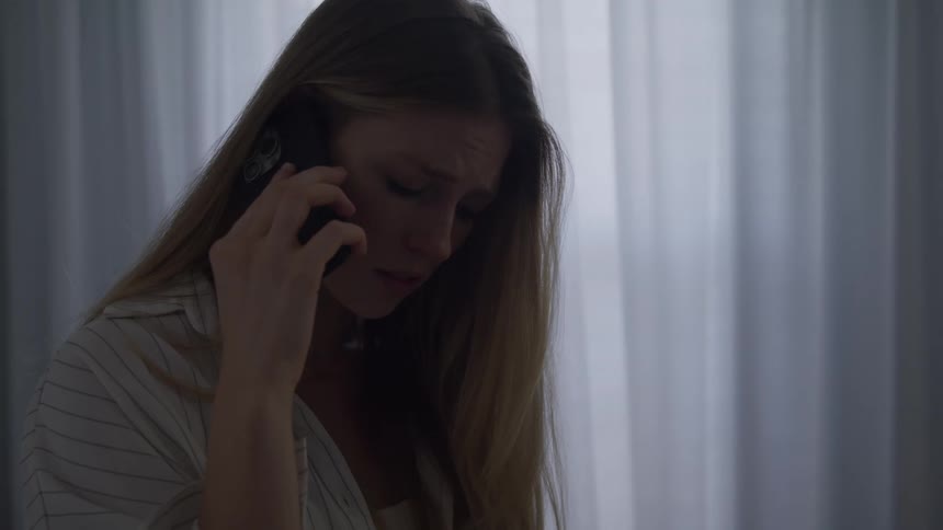 Woman frowns and looks downcast while talking on her smartphone near a curtained window