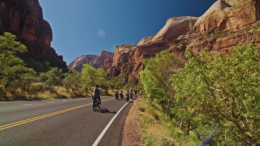 A group of cyclists rides through a sunny canyon and the view sweeps upward to reveal majestic red rock cliffs against a deep blue sky