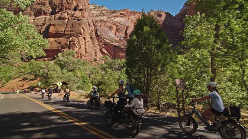 A group of cyclists including men women and children on bicycles and electric bikes ride down a paved road next to towering red sandstone cliffs and lush green trees on a bright sunny day