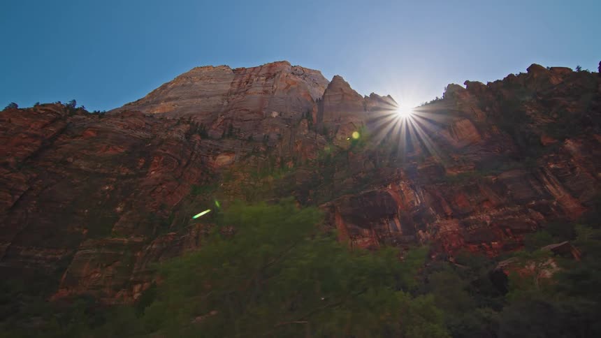 A group of cyclists including a parent with a child ride along a scenic road through a sunlit canyon as a shuttle bus passes and the camera pans up to the majestic cliffs