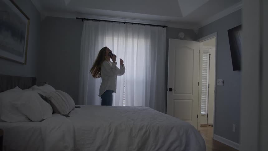 A woman walks around thoughtfully by a window in a softly lit bedroom before answering a call on her cell phone