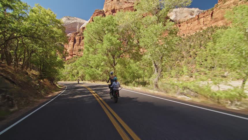 A group of people are bicycling down a paved road surrounded by lush green trees and towering red rock cliffs that suggest a location like zion national park the camera follows two riders one of whom has a child on the back and is pointing out the scene