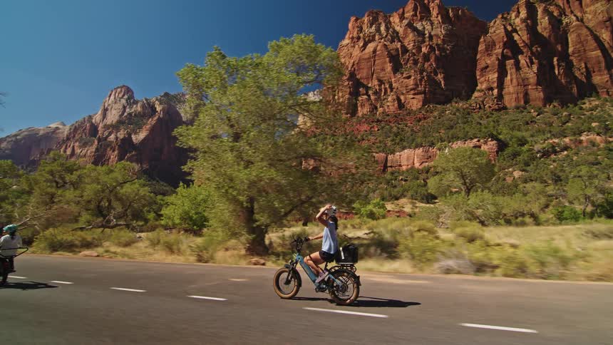 A woman wearing a white helmet and blue shirt rides a thick tire electric bike past towering red sandstone cliffs and vibrant green trees with a line of cyclists following behind her on a sunny road