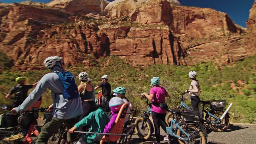 Group of cyclists rides along a road surrounded by canyons under a sunny sky