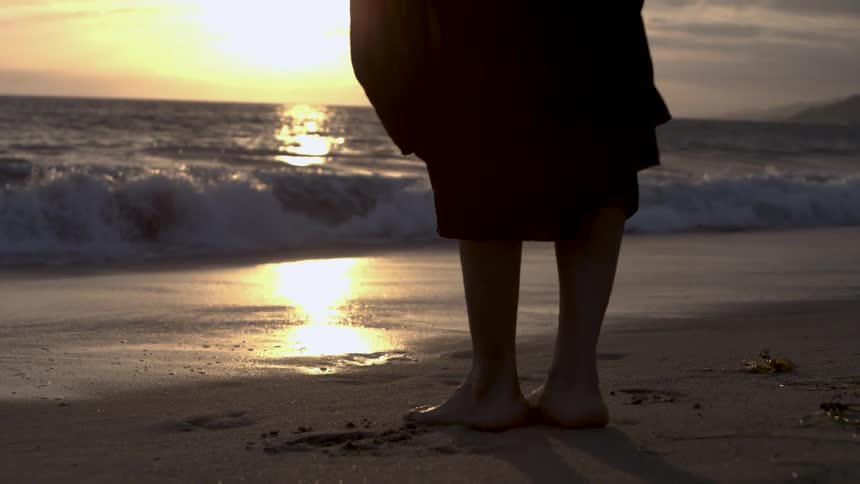 The video captures the bare feet and legs of a person standing on a sandy beach at sunset the sun casts a bright golden reflection on the water as waves crash gently in the background