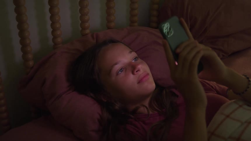 Girl lying in bed looks at her phone under the soft room light