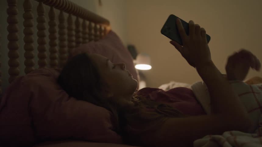 A teenager is laying in bed texting on the phone then puts it down and rolls over