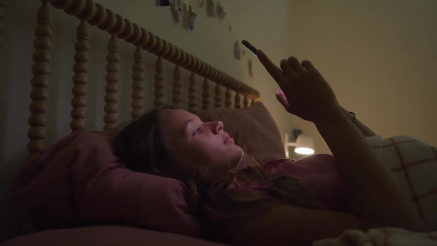 Girl lying in bed uses her phone under the soft light of a bedside lamp