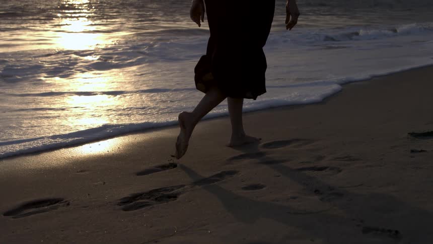 Woman walks barefoot in the sand at the beach leaving footprints at the edge of the waves