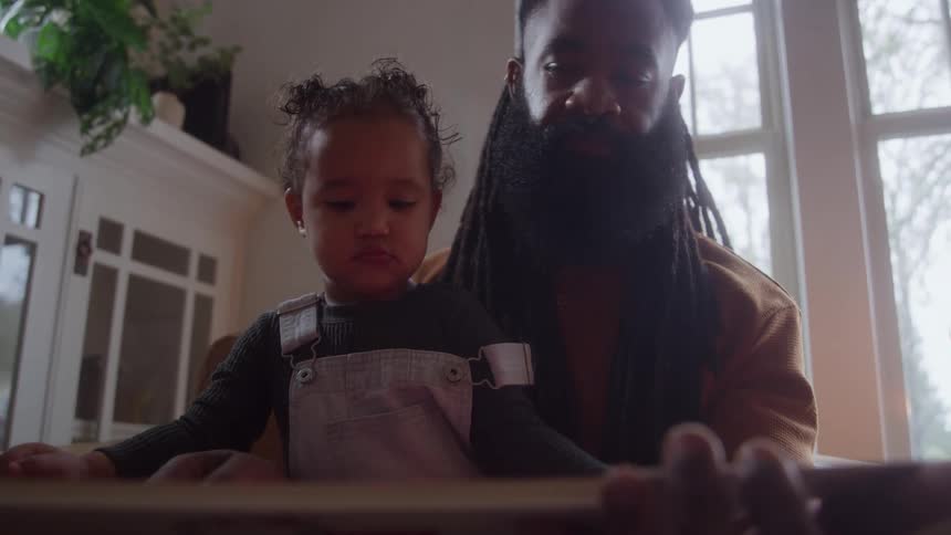 A man with a long beard is seated on a couch with a small child in overalls nestled in his lap they are looking down together at a wooden toy or book in front of them in a bright indoor setting