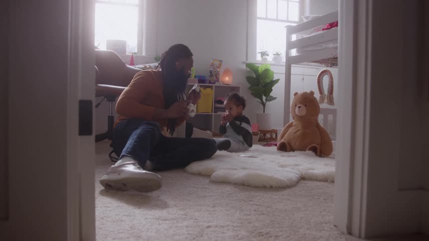 A father and son sit on the floor playing together in a cozy sunlit nursery