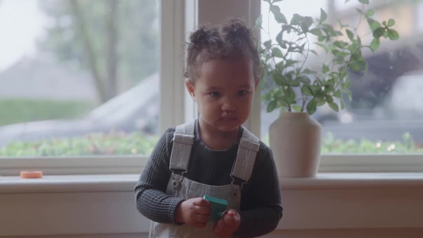 A young child wearing light beige overalls and a dark sweater plays with small colored blocks near a large window looking curiously around