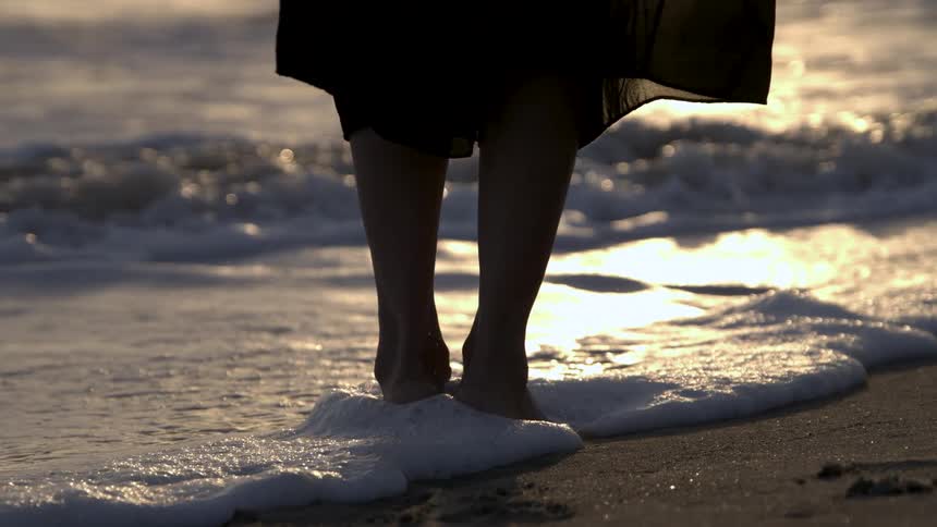 A low close view shows a womans bare legs standing at the edge of the ocean wearing a black dress as the waves wash white foam over her feet under the setting sun
