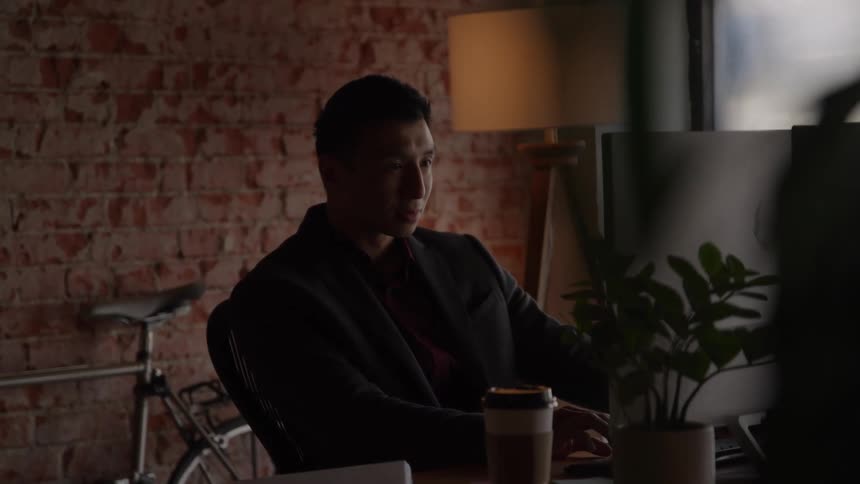 A young man wearing a suit types on a computer while sitting at a desk inside an office
