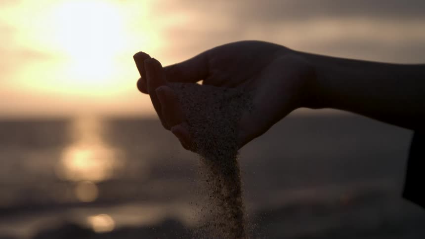 Sand falls from the hand of a woman on the beach as the sun sets on the horizon