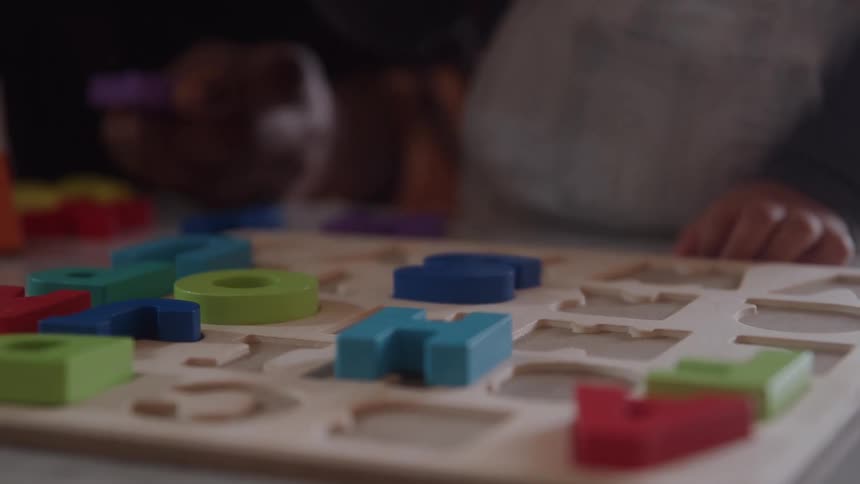 A close view shows a childs hand attempting to fit colorful wooden letter and number puzzle pieces into a matching wooden board on a table in a dimly lit indoor setting