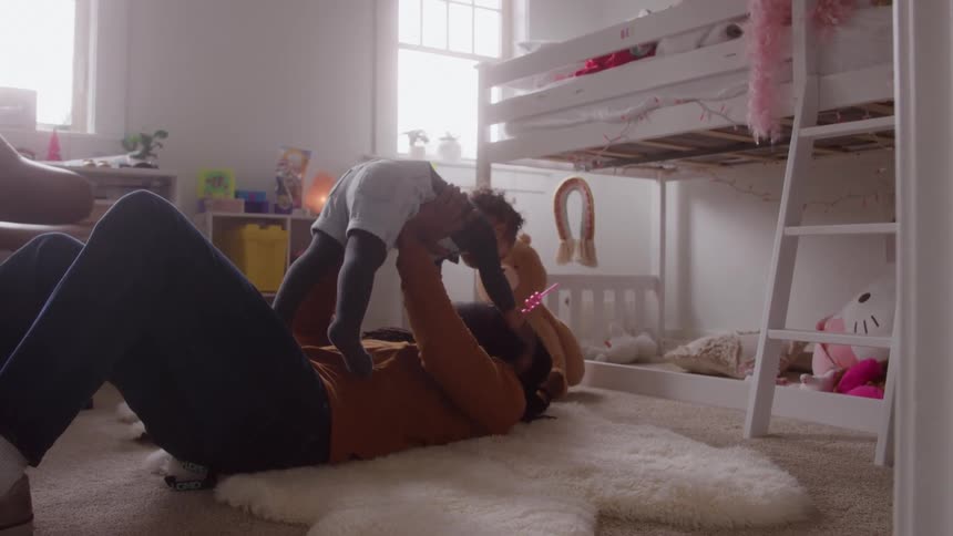 A father lies on a rug in a childs bedroom at midday joyfully tossing his daughter into the air and catching her as sunlight filters through the room