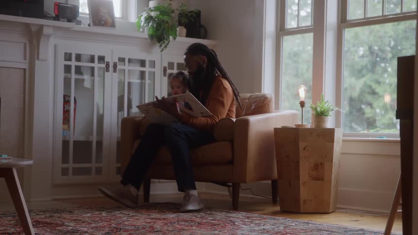The video shows a man with dreadlocks sitting comfortably in a brown leather armchair holding a small child on his lapthey are looking at a book together in a welllit living room with large windows and a decorative builtin cabinet