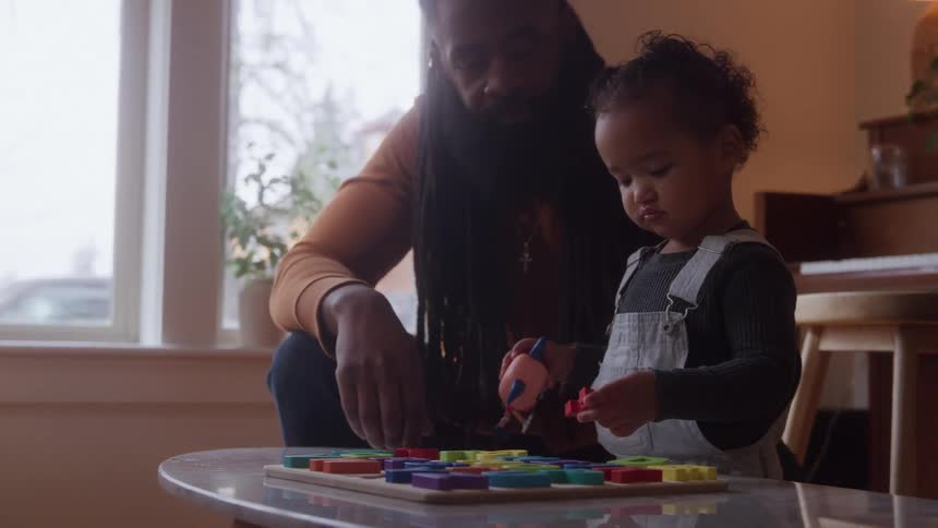 The father is watching his daughter play with blocks