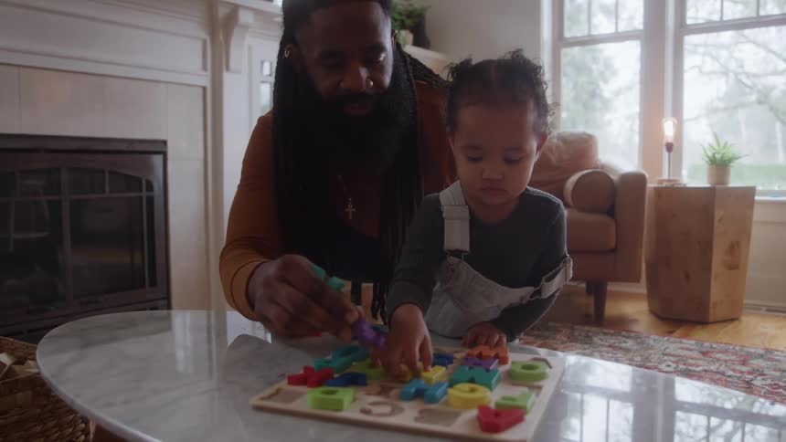 A young man in his living room plays with his toddler daughter helping her arrange letter toys piece by piece at midday