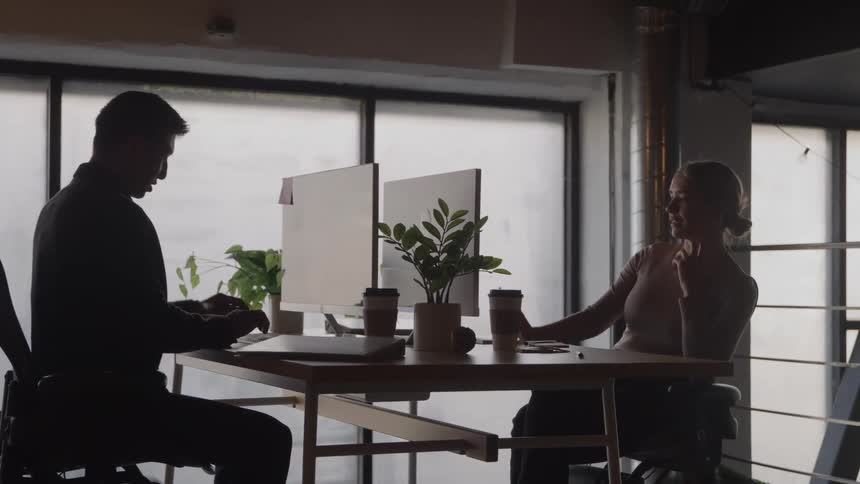 Two co workers conversing while working with desktop computers at a table in an office during daytime