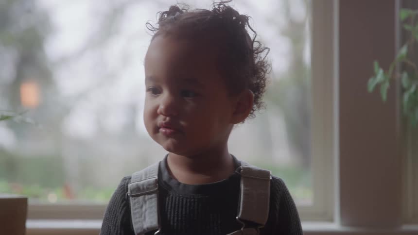 The video is a closeup of a young child with curly hair and suspenders standing near a window the child is looking offcamera with a pensive or serious expression