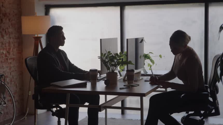 Young male and young female writing and typing on computers on a desk in an office space indoors during day time