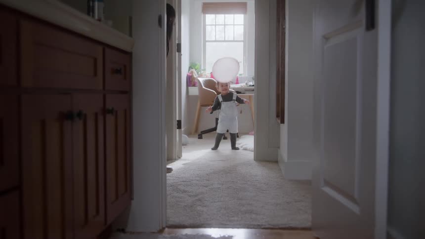 A toddler in overalls walks down a bright carpeted hallway holding a pale balloon that a woman tossed