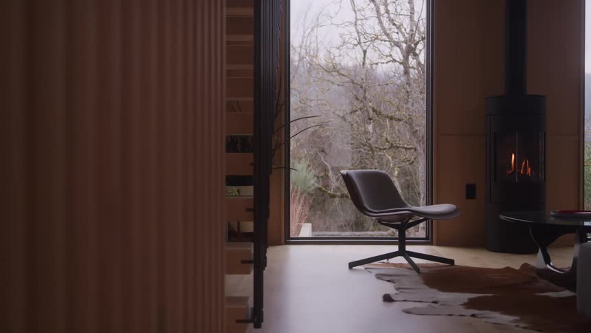 A modern interior room with wood paneling features a gray swivel chair a black standing fireplace and a cow hide rug next to a large floor to ceiling window overlooking a dull green forest