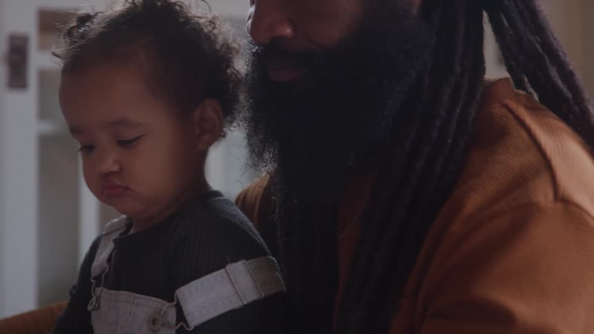 A father sits indoors at midday with his child on his lap gently opening a box in front of her as sunlight filters through the room