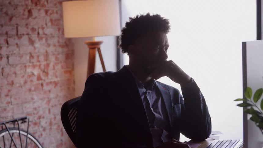 A silhouette of a thoughtful man wearing a suit sits at a desk in an industrial office with a brick wall resting his chin on his hand while looking at a computer screen