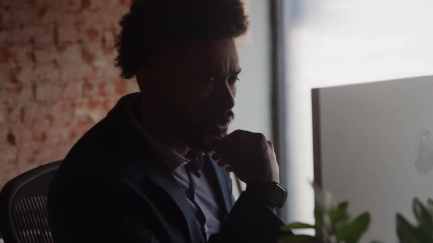 A silhouetted man wearing a suit sits at a desk in an industrial office with a brick wall resting his chin on his hand while intently looking at a computer monitor