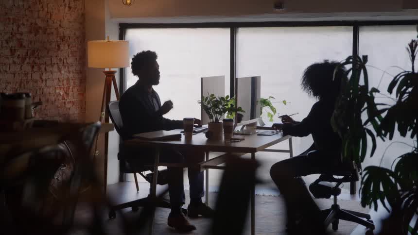A dimly lit interior shows the silhouettes of a man and a woman sitting opposite each other at a desk working on computers near a large bright window and a standing lamp against a brick wall