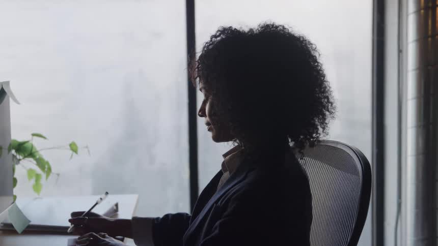 A woman sitting at a desk writes a note a pad of paper with a pen