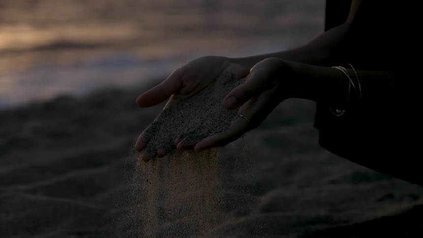 The video is a closeup shot of a persons hands as sand slowly trickles through their fingers onto the beach the scene is set at sunset or dusk on a beach with the gentle light catching the falling grains of sand