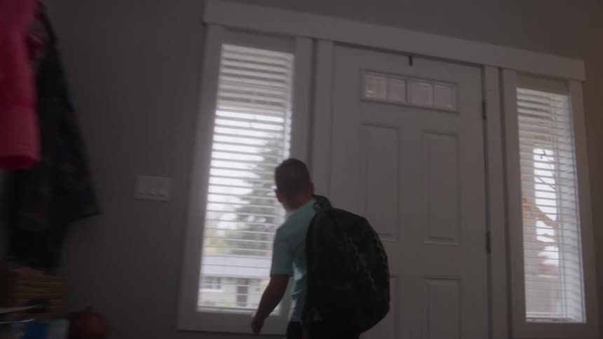 A child wearing a backpack walks from a dimly lit hallway toward the brightly lit front door of a home the child opens the door which has a decorative wreath and then steps outside