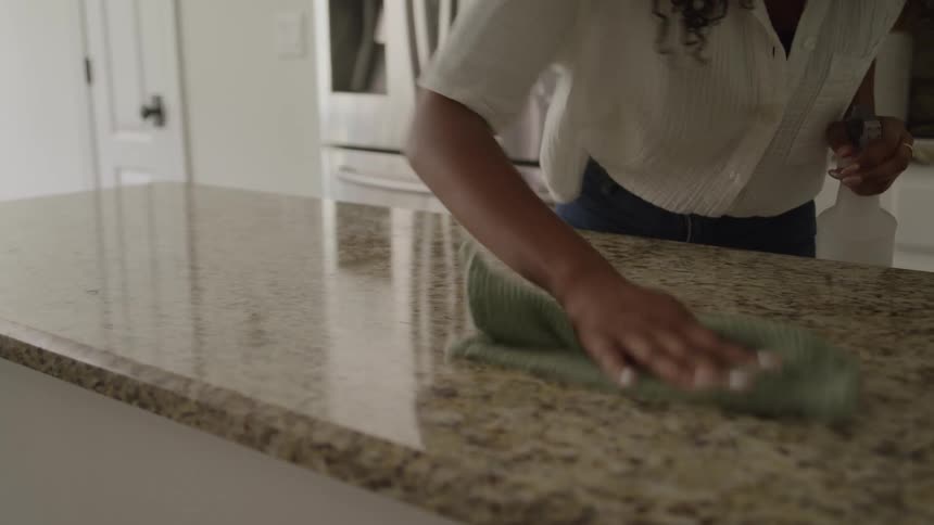 A person in a kitchen is shown cleaning a granite countertop they spray a cleaner onto the surface from a white bottle and wipe it down with a dark green cloth