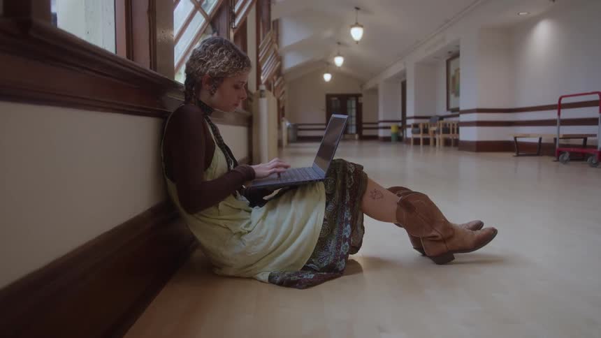 A woman sits on the floor leaning against a wood paneled wall the woman holds a laptop in her lap and types on the keyboard a long hallway extends into the background windows run along the wall behind the woman the illumination is overhead lights
