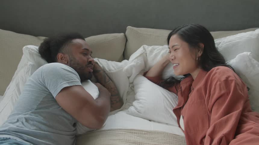 Black man and biracial woman lying down in a bed talking and smiling indoors