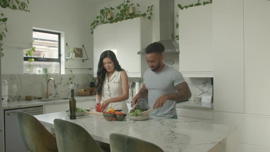 Young black man and young biracial woman standing up in a kitchen preparing vegetables indoors during day time