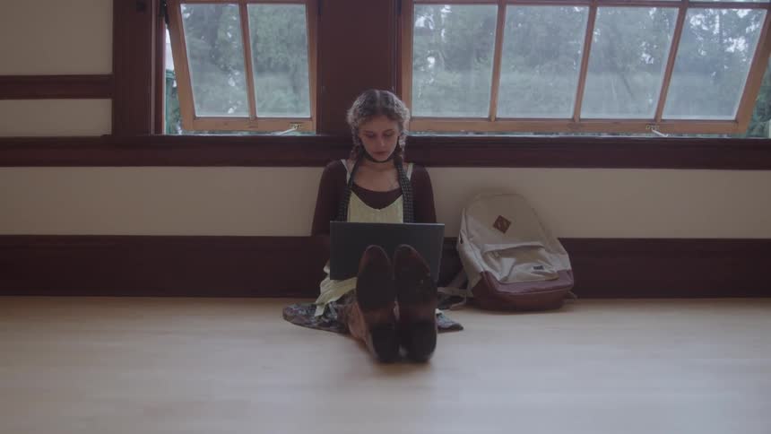 Young teenager girl sitting down on a wooden floor on a corridor using a laptop computer indoors during day time