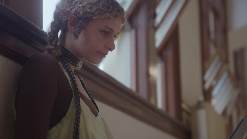 The video is a closeup of a young woman with curly light brown hair looking down possibly at a book or device she is indoors in a large bright space with architectural wood banisters and railings visible in the background