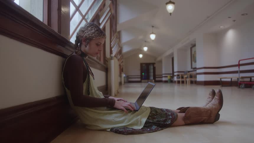 Woman sitting on the floor of a hallway types on a laptop computer