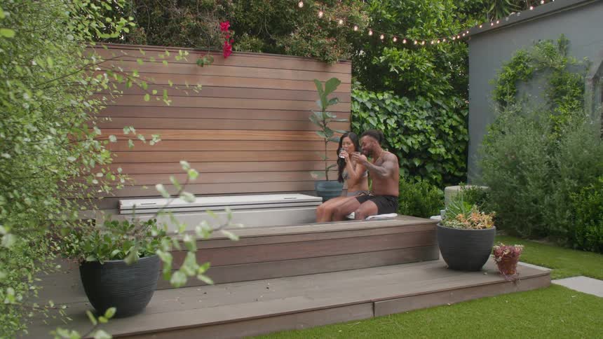 The video features a couple relaxing in a wooden outdoor hot tub on a sunny day in a lush fencedin backyard they are talking and smiling at each other conveying a sense of enjoyment and intimacy