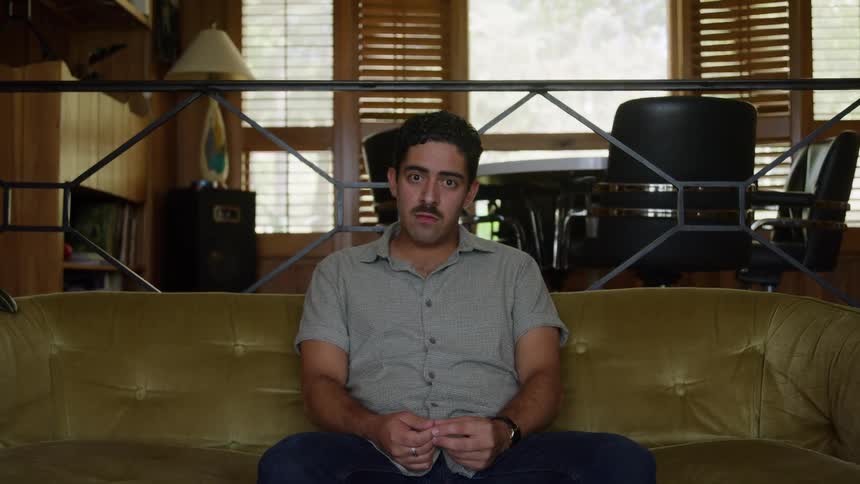A serious looking man with dark hair and a beard sits in the center of a gold velvet tufted couch wearing a gray collared shirt and jeans in a brightly lit midcentury modern room
